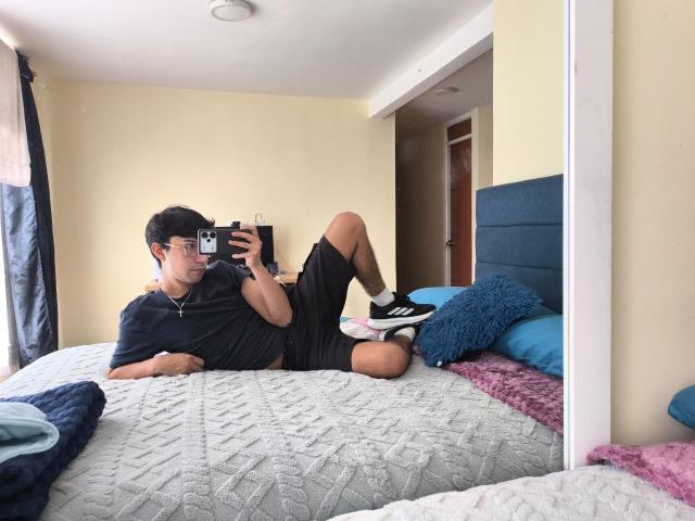 OliverHot69, lying on a bed, takes a mirror selfie. Wearing a black t-shirt, black shorts, and black-and-white sneakers, OliverHot69 holds a black smartphone in his right hand. He has short black hair and wears glasses. The bed has a textured gray quilt, blue and purple pillows, and a blue headboard. OliverHot69's left leg is bent at the knee, and his right leg is stretched out. The room has beige walls and a white ceiling. The mirror shows OliverHot69's reflection.
