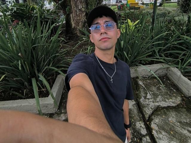 OliverHot69 selfie, wearing a black cap, reflective sunglasses, dark navy t-shirt, cross necklace, black wristband, short hair, fair skin, small nose, slight smile, outstretched arm, green plant background.