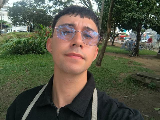 OliverHot69 hat kurze, schwarze Haare, trägt reflektierende Sonnenbrillen und ein schwarzes, vorne zu öffnendes Hemd mit grauen Trägern. Er steht in einem grünen Park mit Bäumen und einem Fahrrad..