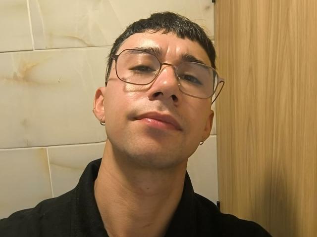 OliverHot69 con gafas, cabello oscuro corto, ligera sonrisa, camisa negra, aretes pequeños, piel clara, ligero vello facial, puerta de madera a la derecha..