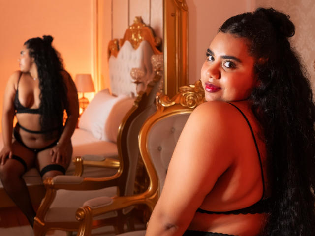 LisaTobon with long black curly hair, wearing black lingerie, sits on a gold chair, looking back at her reflection in a mirror, showing her big ass and small tits.