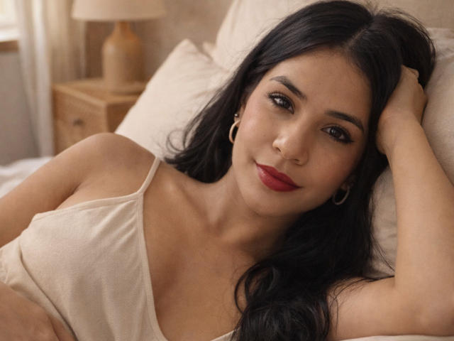 AmaiaCooper with long black hair, lying on a bed, wearing a beige tank top, red lipstick, and hoop earrings, looking at the camera with a subtle smile.