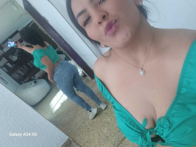 ScarlettOxford with puffy lips, green off-shoulder top, showing big tits, blue jeans, and white sneakers, reflected in mirror, standing with ass facing camera.