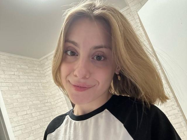 KerryColver with blonde hair, green eyes, nose ring, black and white raglan shirt, smiling, small earrings, light skin, soft makeup, slightly messy hair, subtle pink lips.