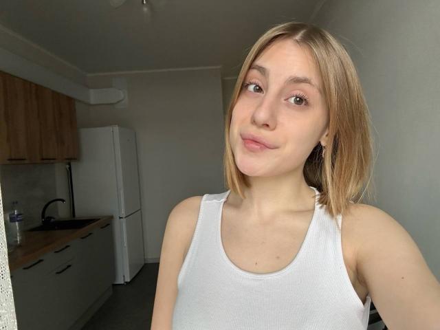 KerryColver, blonde, white tank top, small tits, fair skin, light brown eyes, slight smile, small nose, short hair, kitchen background, white fridge, wooden cabinets, gray floor.