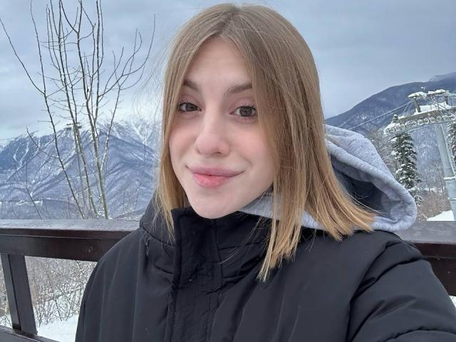 KerryColver with medium-length blonde hair, wearing a black puffy coat, has fair skin and a slight smile, standing outdoors with a snowy mountain background and a cloudy sky.