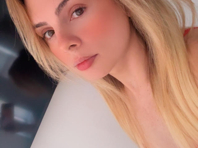 BellaCampos has blonde hair, pink blush, and pink lips, with a slightly pouty expression, looking to the side. Her tits are not visible, just her face.
