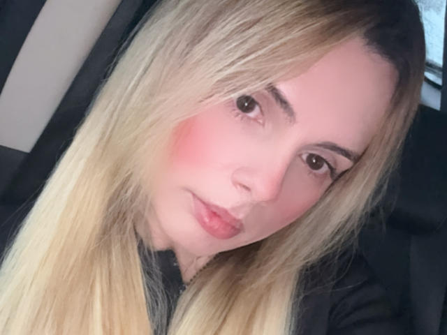 BellaCampos has blonde hair, pink cheeks, and pouty pink lips. She's looking at the camera with brown eyes and a slight smile, wearing a black top.