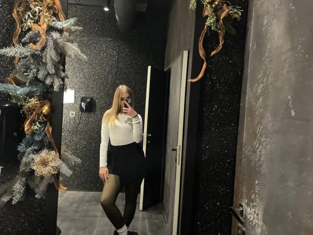 AlinaLemon with blonde hair, white long-sleeve top, black high-waisted skirt, green tights, and white socks, taking a mirror selfie in a dark, textured room.