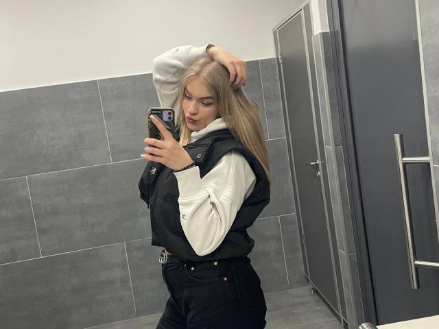 AlinaLemon with blonde hair, white hoodie, black vest, and black pants, taking a mirror selfie, hand in hair, looking at phone, grey tiled bathroom, medium ass.
