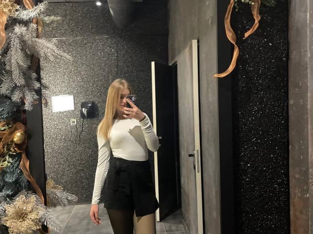 AlinaLemon with blonde hair, white long-sleeve top, black high-waist shorts, and black tights, taking a mirror selfie, showing her ass, in a dark, textured hallway.
