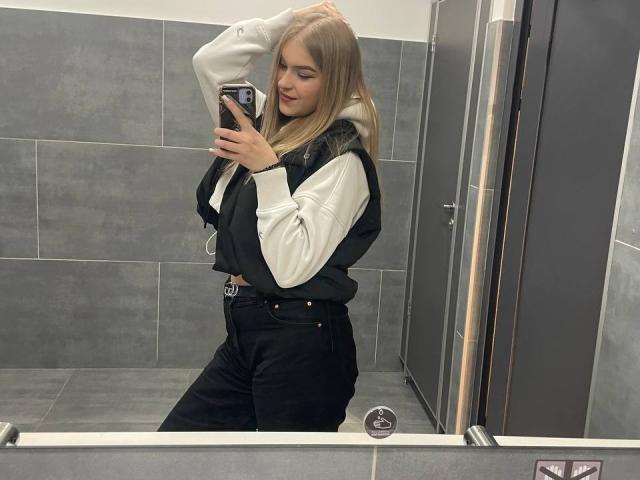 AlinaLemon with blonde hair, white hoodie, black vest, and black pants, taking a selfie in a bathroom mirror, showing her ass and tits, holding a phone.