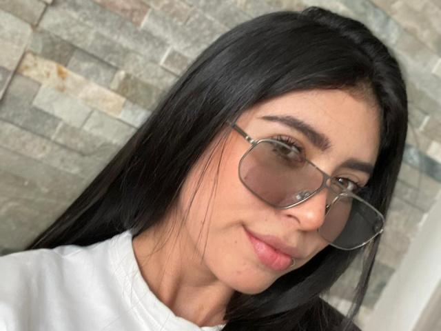 MiluSky with long black hair, wearing glasses and a white shirt, has a slight smile, showing pink lips and brown eyes. Her nose is small and she's cute.