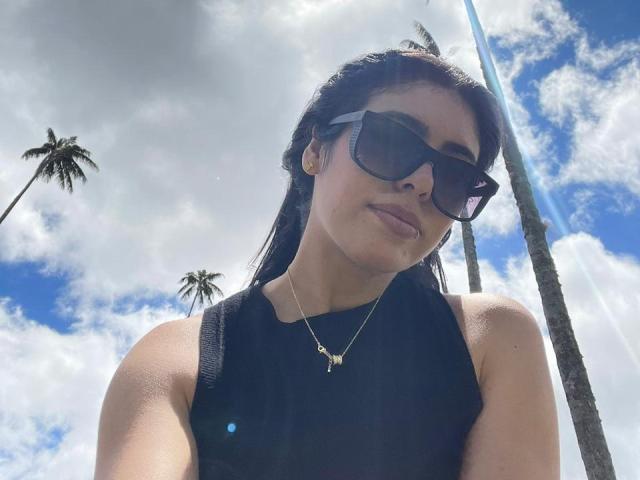 MiluSky con gafas de sol oscuras, top negro sin mangas, collar dorado y busto medio, de pie al aire libre, con palmeras en el fondo, un día soleado, con la cabeza ligeramente inclinada, mirando hacia abajo..