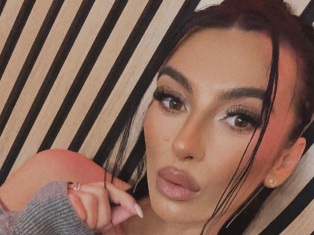 KinkyAmelie has dark hair with pink highlights, thick eyebrows, long eyelashes, and full pink lips. She's wearing a gray sleeve and has her finger near her mouth.