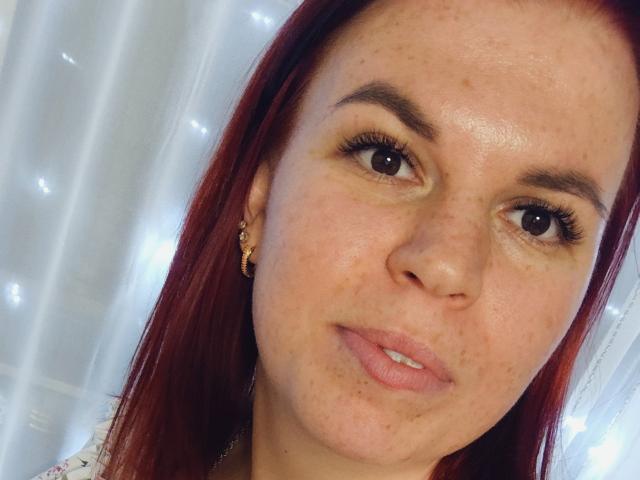 VeronikaLune has red hair, brown eyes, and freckles on her face. She's wearing gold earrings and has a slight smile with pink lipstick on her "pussy" (lips).