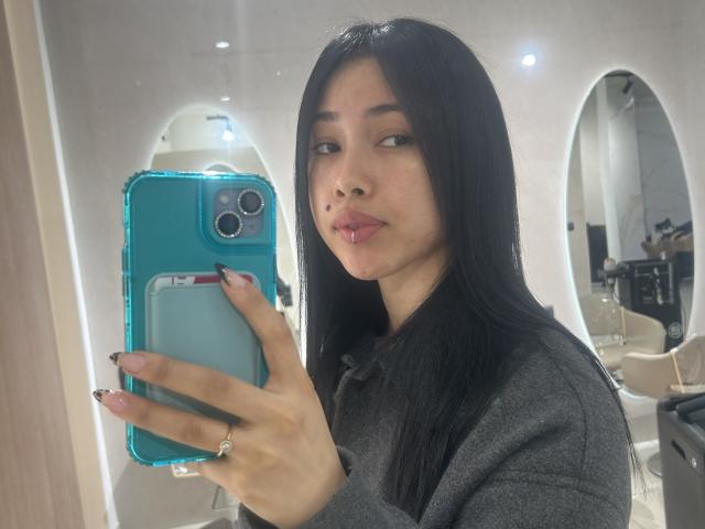 NashiroSakuraii with long black hair, wearing a gray sweater, holding a blue phone, pouting lips, looking in mirror, wearing a gold ring on her right hand.