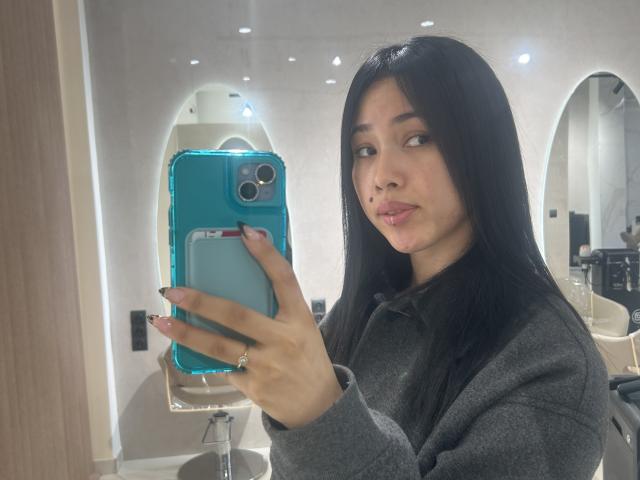 NashiroSakuraii with black hair, wearing a gray sweater, taking a selfie in a mirror with a blue phone, showing her small, round, mochito-shaped nose, and pink lips.