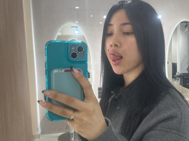 NashiroSakuraii with long black hair, tongue out, holding a blue phone, wearing a gray shirt, ring on finger, in a mirror selfie. Her nails are long and painted.