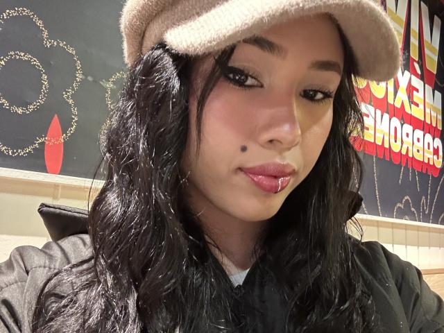 NashiroSakuraii has long black wavy hair, a brown flat cap, and a black jacket, with a small beauty mark on her right cheek, and glossy pink lips.