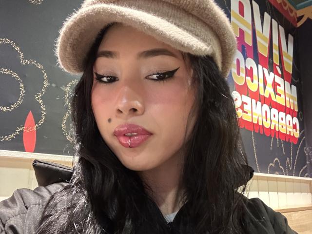NashiroSakuraii with dark wavy hair, pink glossy lips, black eyeliner wings, beige wool hat, black jacket, small beauty mark on left cheek, slightly puckered lips.