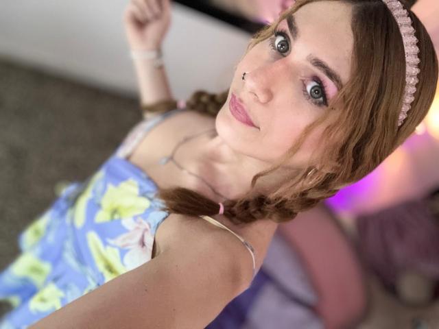 AndyDupont with light brown braids, pink headband, nose ring, blue floral dress, pink lipstick, and blue eyes, looking up at camera with a slight smile. Boobs visible.
