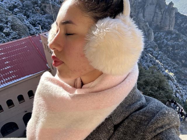 NashiroSakuraii with white fluffy earmuffs, pink scarf, and grey herringbone coat, eyes closed, pink lipstick, and soft light skin, standing outdoors with rocky mountains and red roof in background.