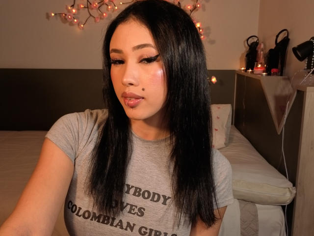NashiroSakuraii with long black hair, wearing a gray t-shirt, has pouty pink lips, bold eyeliner, and a small beauty mark on her right cheek. Warm bedroom lights in background.