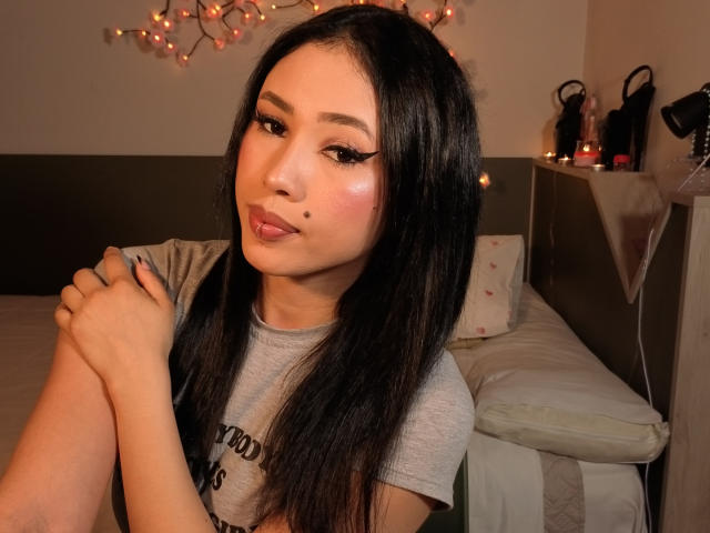 NashiroSakuraii with long black hair, pouty pink lips, and a beauty mark on her right cheek, wearing a gray shirt, hands folded, in a warmly lit room.