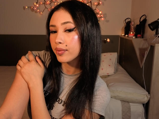 NashiroSakuraii with long black hair, pink eyeliner, and glossy pink lips, wearing a gray shirt, hands together, a small mole on her right cheek, in a softly lit room.