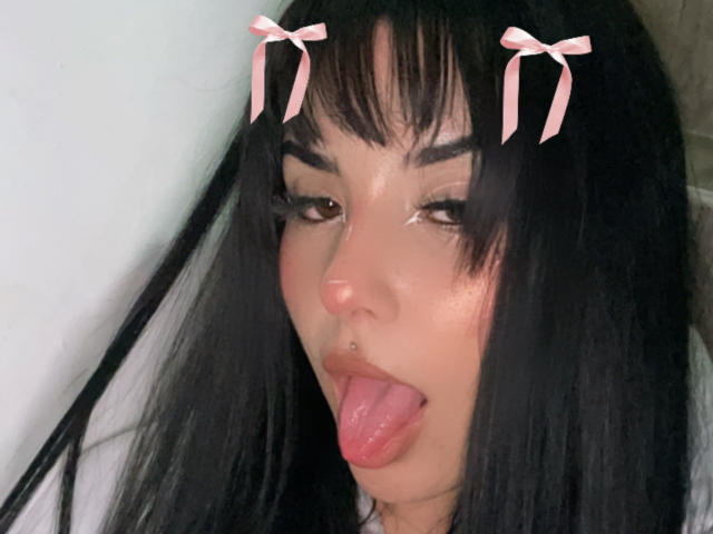 GraceClarke has long black hair with pink bows, brown eyes, a nose piercing, and her tongue out. She's winking and looking slightly up. Her lips are slightly parted.