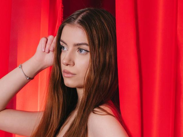EricaDiaz with long brown hair, blue eyes, and light skin, holding her head with a silver bracelet, looking serious, standing behind bright red curtains.
