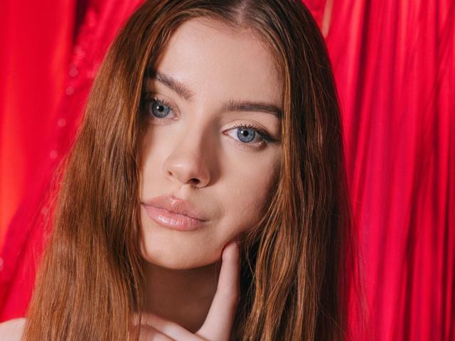 EricaDiaz's face is centered in the image, with her right hand gently touching her chin. She has long, straight brown hair, blue eyes, and a light skin tone. Her expression is calm and slightly pouty. The red background is textured and bright, contrasting with her skin and hair. The lighting is even, highlighting her facial features and the subtle sheen on her lips. The overall composition is balanced, focusing on EricaDiaz's face.