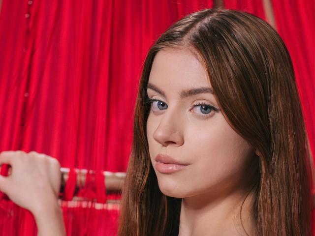 EricaDiaz with long brown hair, blue eyes, and nude pink lips, holding a red curtain rod in front of red curtains, looking directly at the camera with a neutral expression.