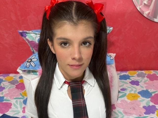RilleyFoyx has long brown pigtails with red bows, wearing a white shirt and red plaid tie, sitting on a colorful floral bed, with a pillow behind her.