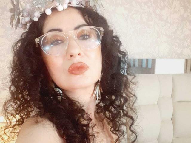 SamanthaAntolin has curly black hair, wearing clear, oversized glasses. She has light skin with a slightly flushed complexion, and is making a pouty expression with her lips. She's wearing a floral headband with white pearls. Her eyebrows are thick and slightly arched. She has dark, well-defined eyebrows and is looking directly at the camera. The background is a textured, light-colored wallpaper. She's positioned in a white, tufted headboard. The image is bright with a warm, soft light. She's wearing a strapless top.