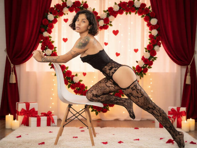 AnneeFoxy with tattoos, black lace bodysuit, and thigh-highs, kneels on a white chair, showing her ass and pussy, in front of a heart-shaped red and white flower wreath.