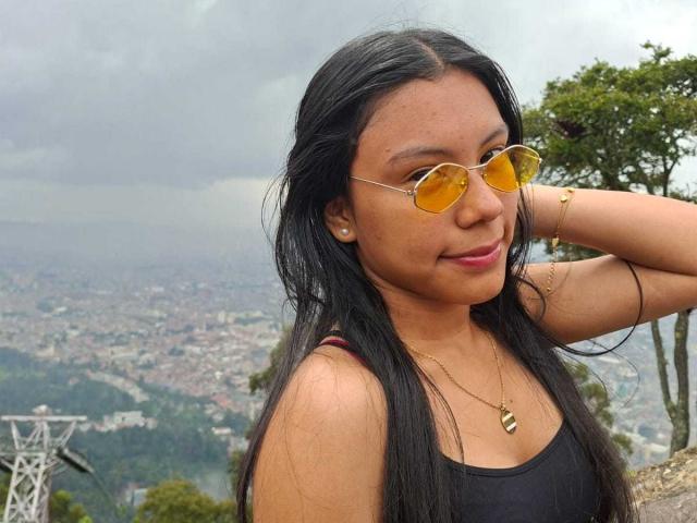 IvyMontaner with long black hair, yellow-tinted aviator sunglasses, black tank top, gold necklace, and bracelet, smiling, hand behind head, overcast sky, city and green hills in background.