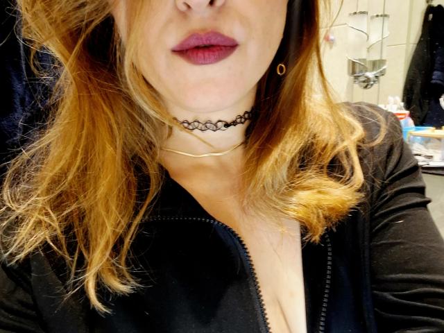 Millfka's face showing a bit of her tits, with dark red lipstick, a black choker, and a gold necklace, wearing a black zip-up top, blonde hair.