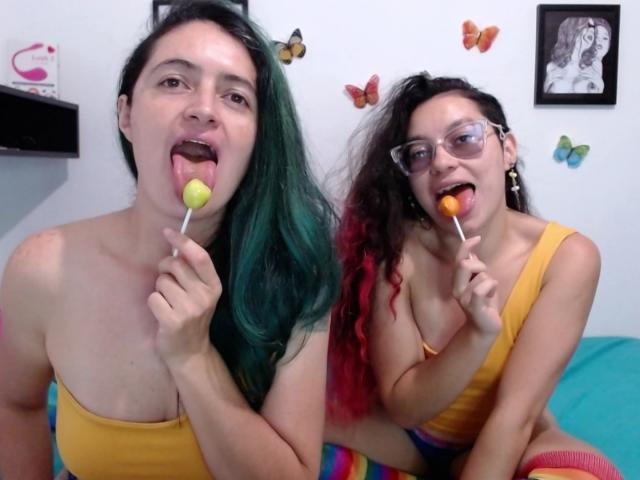 LunaVenus with green-blue hair and yellow tank top, sticking out tongue, holding yellow lollipop. LunaVenus with red-black hair, glasses, yellow tank top, holding orange lollipop.