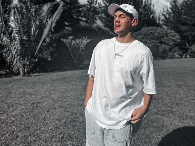 LordThoring, wearing a white cap and shirt, with hands in jeans, smiling, has a necklace and is standing on grass with trees in the background.