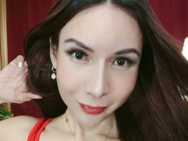 Close-up of MissHotAyaLove. She has long, dark brown hair flowing over her right shoulder, with her right hand gently touching her hair. Her brown eyes are large and framed with thick, black eyebrows. She wears subtle, natural-looking makeup with red lipstick. Her skin is fair and smooth. She has a small, gold, floral earring in her right ear. She's wearing a red strap on her right shoulder, and a thin gold necklace. The red background contrasts with her skin tone and hair color.