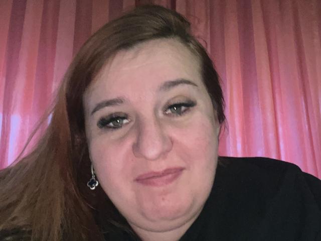 CarolineAva with light skin, brown hair, and dark eyeliner, wearing a black shirt, has a small nose and is smiling slightly with a pinkish background.