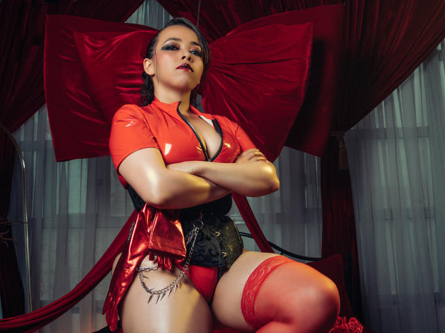 BarbaraLeythonn with crossed arms, red latex top, black corset, red panties, red thigh-highs, and chain on left thigh, sitting against large red bow, looking confident.