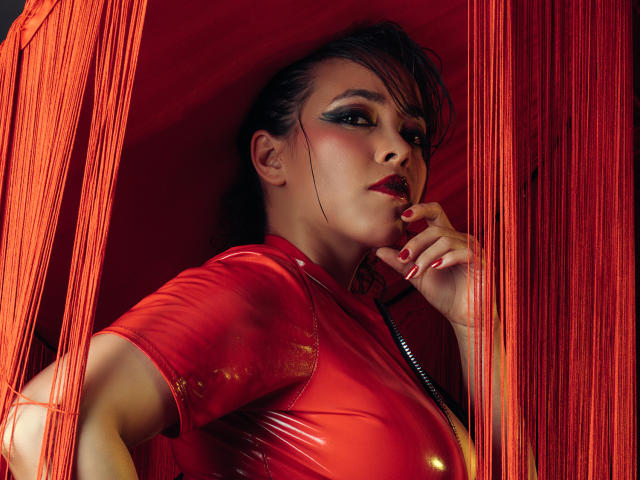 Barbara Leythonn con rossetto rosso, trucco occhi scuro e unghie rosse, indossa una maglia rossa lucida, con le dita vicine alla bocca, e tende di un sipario rosso su entrambi i lati..