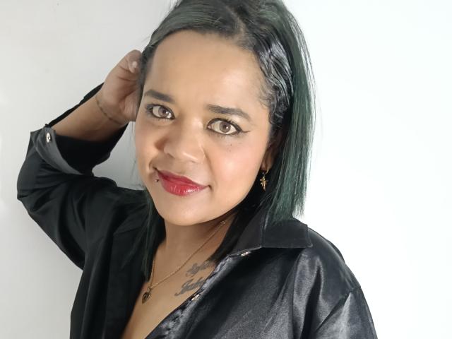 SamantaLevis with dark green hair, black shirt, red lipstick, tattoo on chest, hand in hair, smiling, brown eyes, gold earrings, light skin, medium build, attractive.