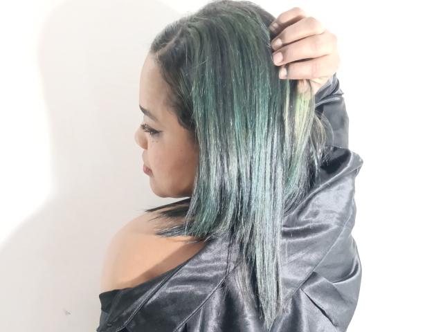 SamantaLevis with green-tinted, straight bob, holding her hair up, wearing a black leather off-shoulder top, showing her bare shoulder and side profile.