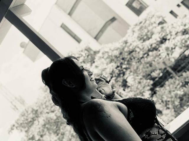 NikolUnlimit with dark hair in a bun, wearing a strapless top, holding a cigarette to her mouth, looking up, black and white, subtle tattoo on her shoulder.
