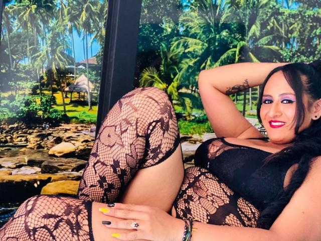 NikolUnlimit with big tits, wearing black lace fishnet lingerie, lying down, smiling with bright pink lipstick, black hair, yellow nails, tattoo on arm, and a bracelet.