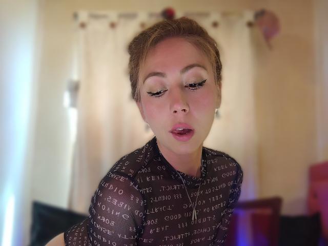 DouceKhrystine with her eyes half-closed, wearing a sheer black top with white text, has pink lipstick and subtle makeup, looking slightly pouty in a soft-lit room.